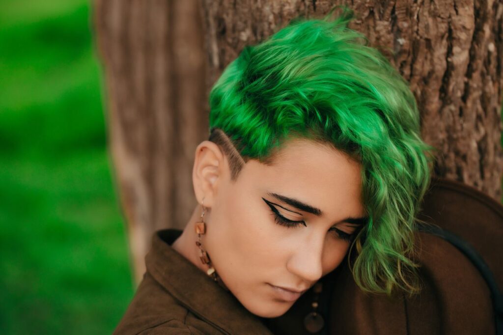 Close-up portrait of a woman with striking green hair posing outdoors against a tree.