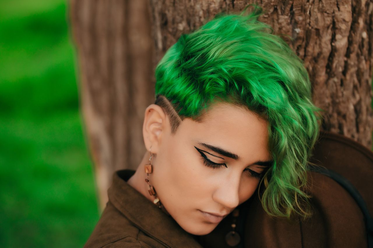 Close-up portrait of a woman with striking green hair posing outdoors against a tree.