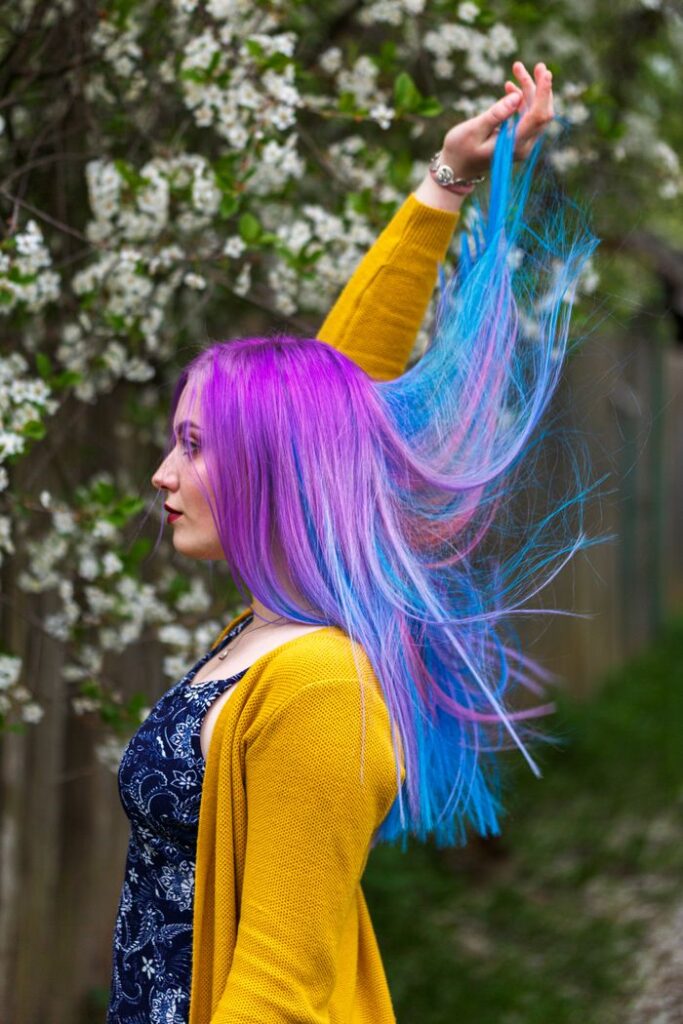 Woman with purple and blue hair in yellow sweater amidst blooming spring flowers.