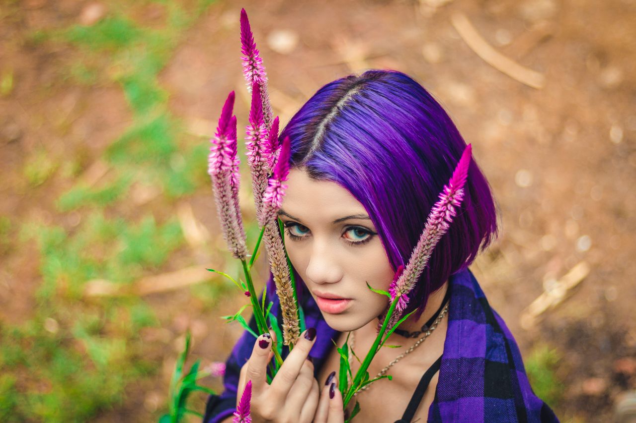Close-up portrait of a woman with purple hair and vibrant flowers, outdoors.