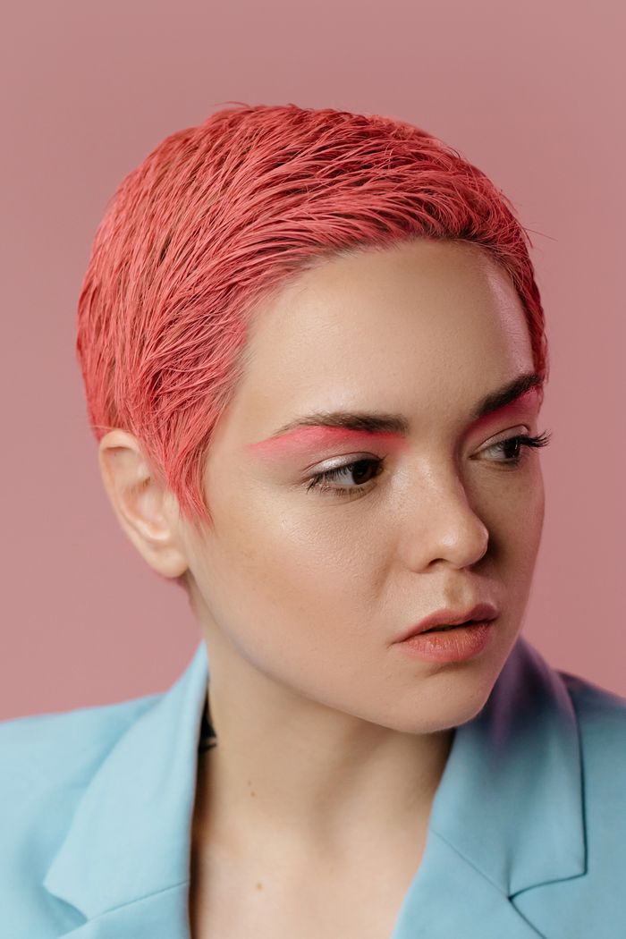 Striking headshot of a young woman with pink hair and blue jacket on pink background.
